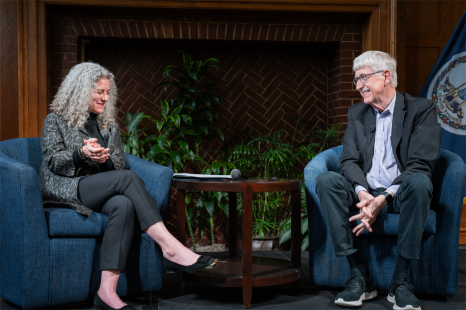 Two people engaged in a conversation sit in blue chairs on a stage with a table between them